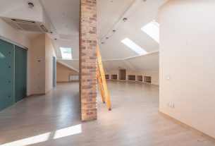 An empty modern attic room with a wooden loft ladder placed against a brick pillar and skylights above.