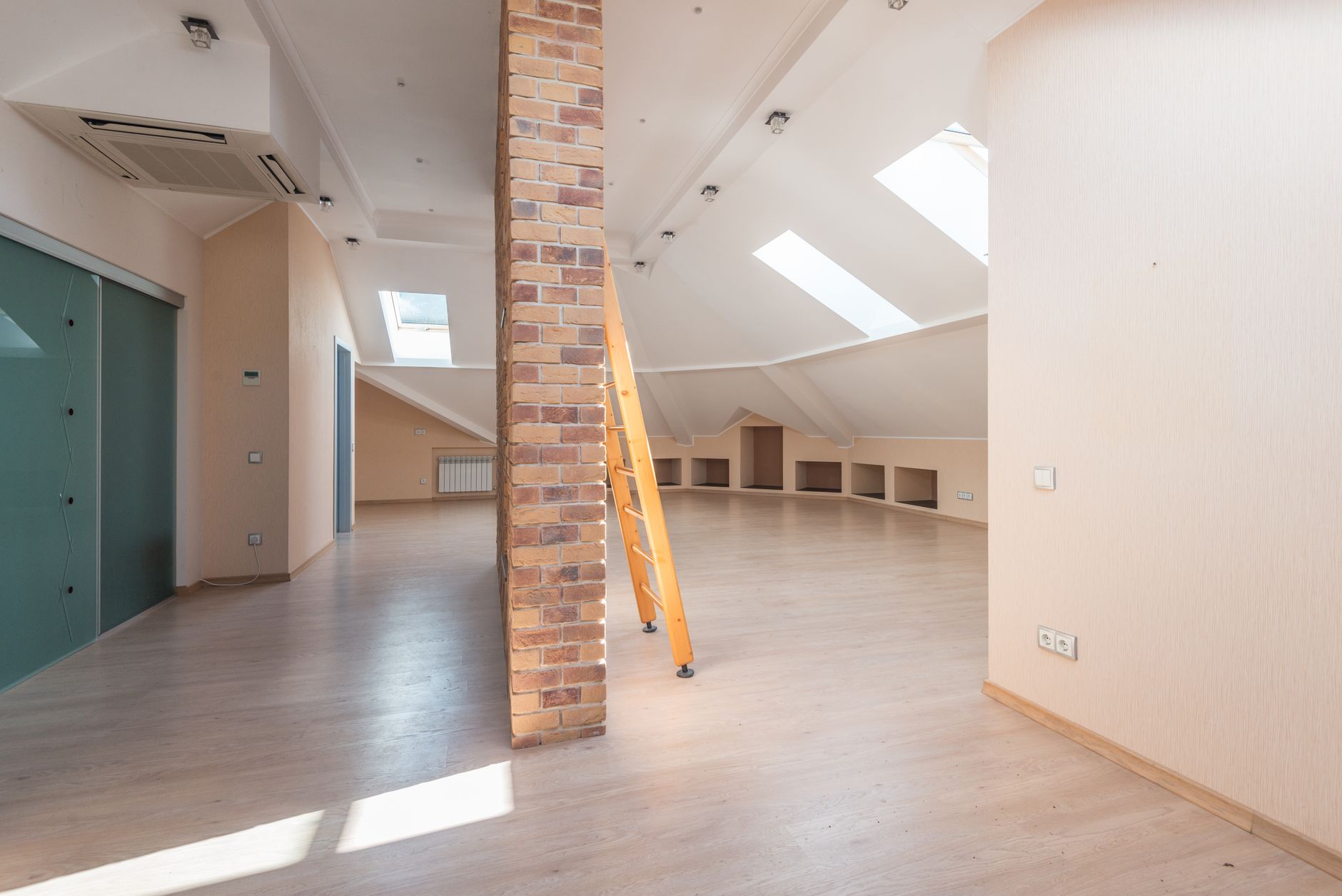 An empty modern attic room with a wooden loft ladder placed against a brick pillar and skylights above.